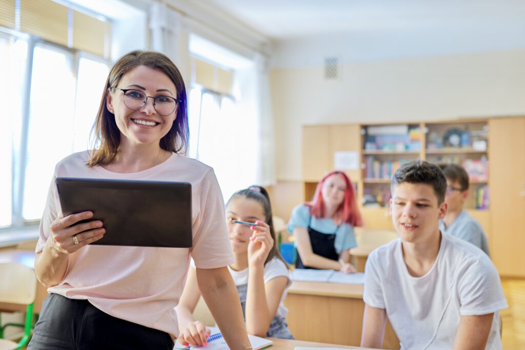 Portrait of female teacher smiling at camera in classroom with teenage students at desks