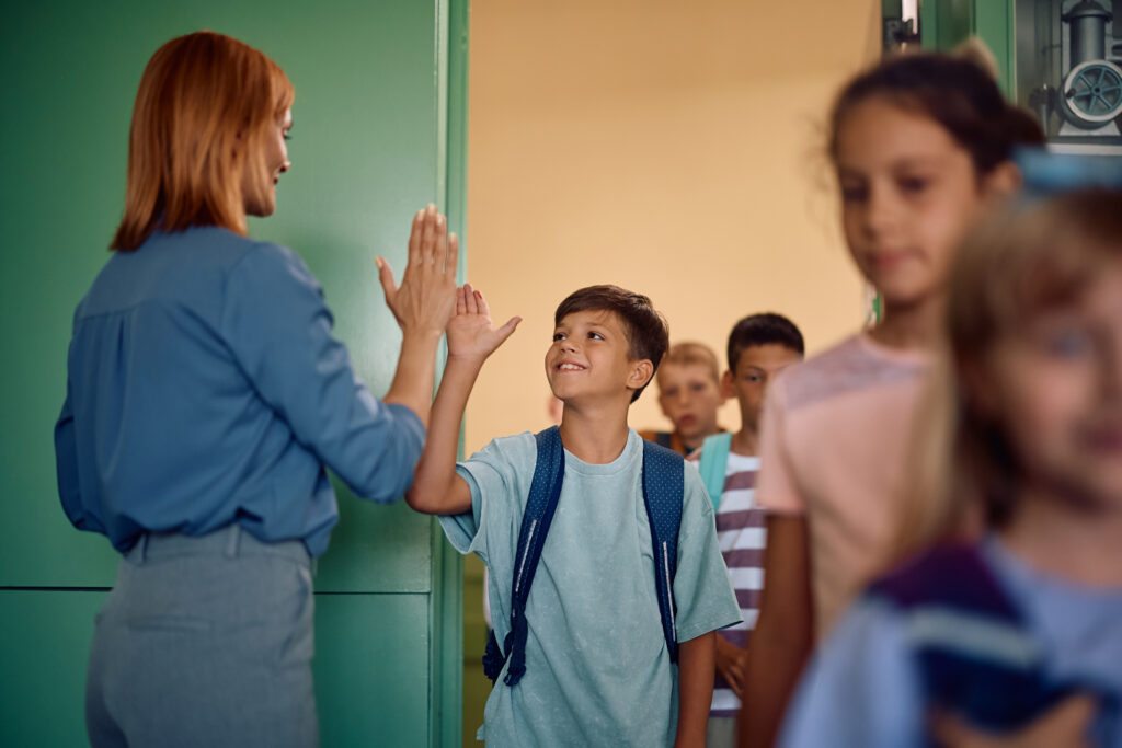 happy boy giving high-five to his teacher while walking into a classroom with his classmates