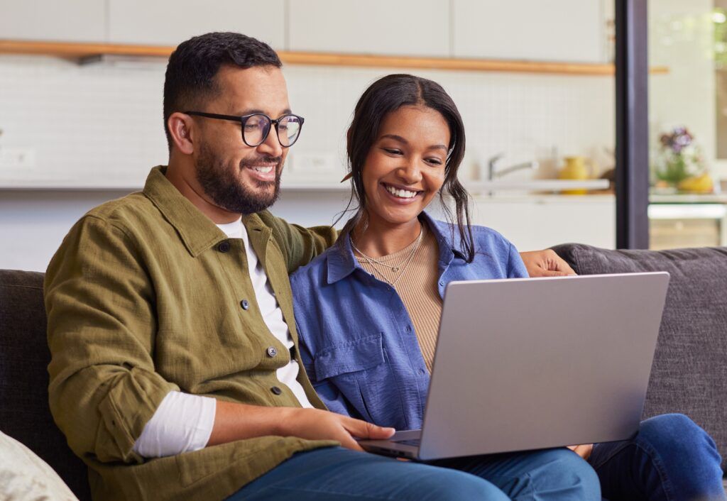 smiling couple sitting on the couch at home on the couch looking at a laptop screen