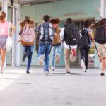 Group of school children wearing backpacks running down a covered outdoor school hallway toward a building entrance, captured from behind during the school day.