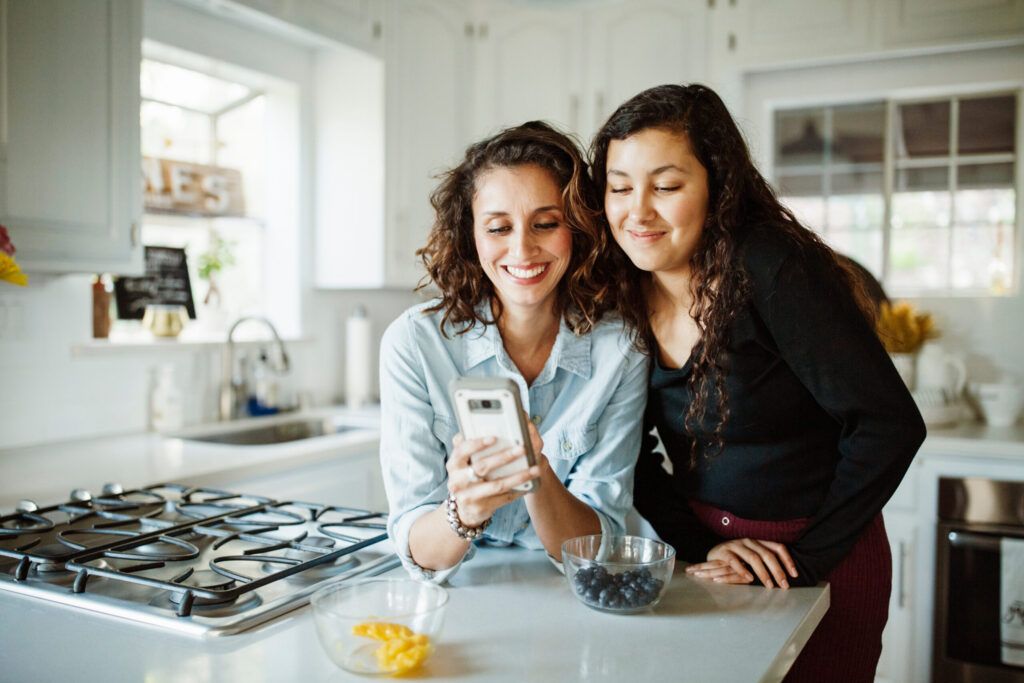 Smiling mother and daughter using mobile phone while standing by kitchen counter at home