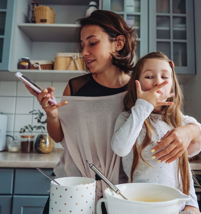 A woman and a young girl are in a kitchen in the middle of baking. The woman is holding a smartphone in one hand while wrapping her other arm around the girl.