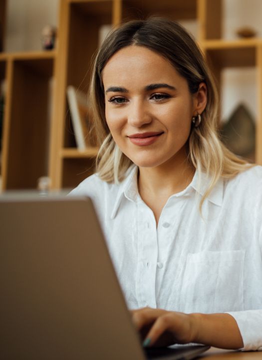 A smiling woman in a white shirt looking at a laptop screen in an office or home setting.