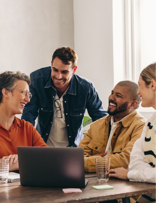 Four coworkers smiling and talking around a laptop during a collaborative meeting in a bright office setting.