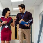 Two school staff members wearing ID badges walk through a hallway while talking, with one holding an open folder and pointing during their discussion.