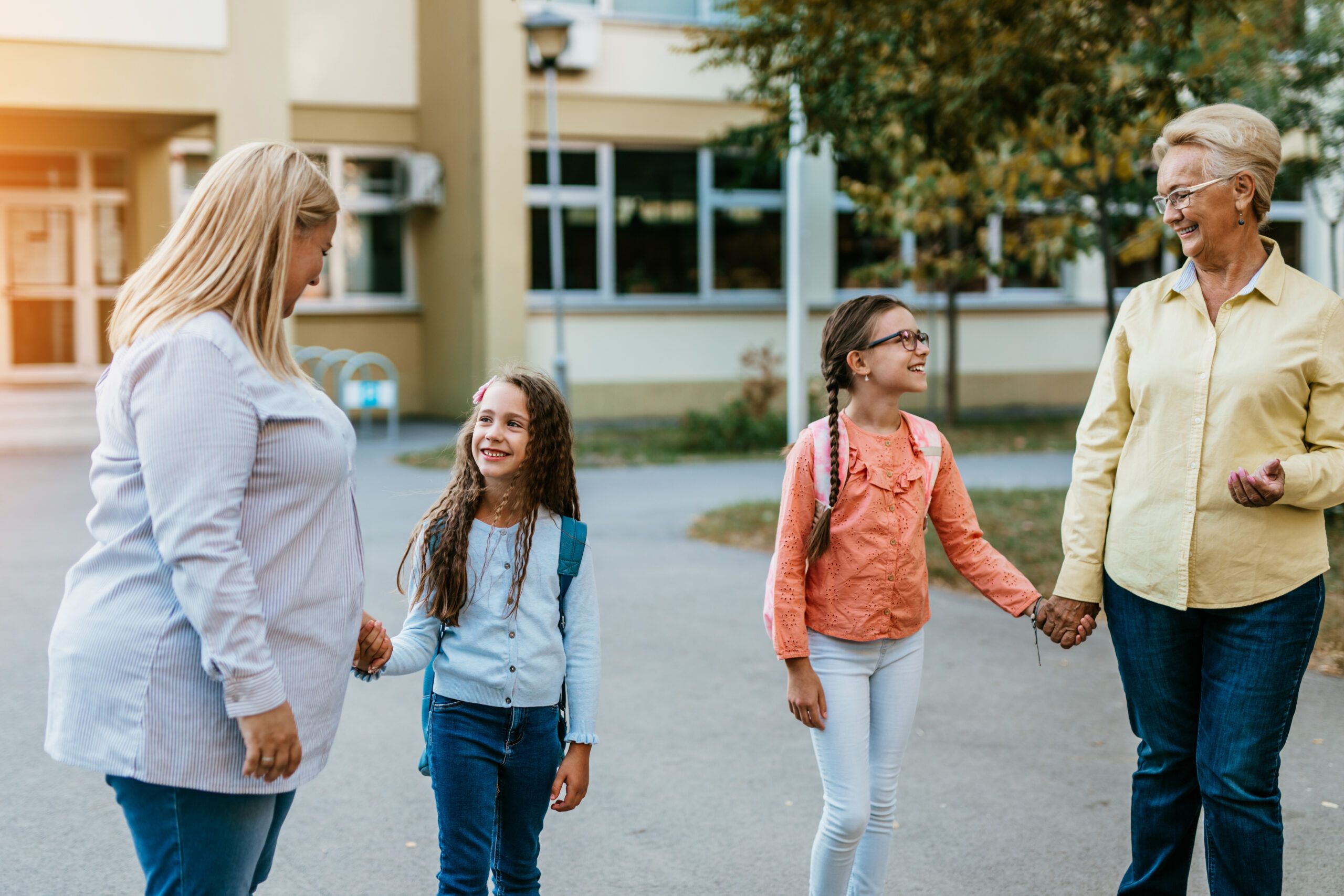 Two adults and two school-age girls walking together outside a school building while holding hands and smiling.